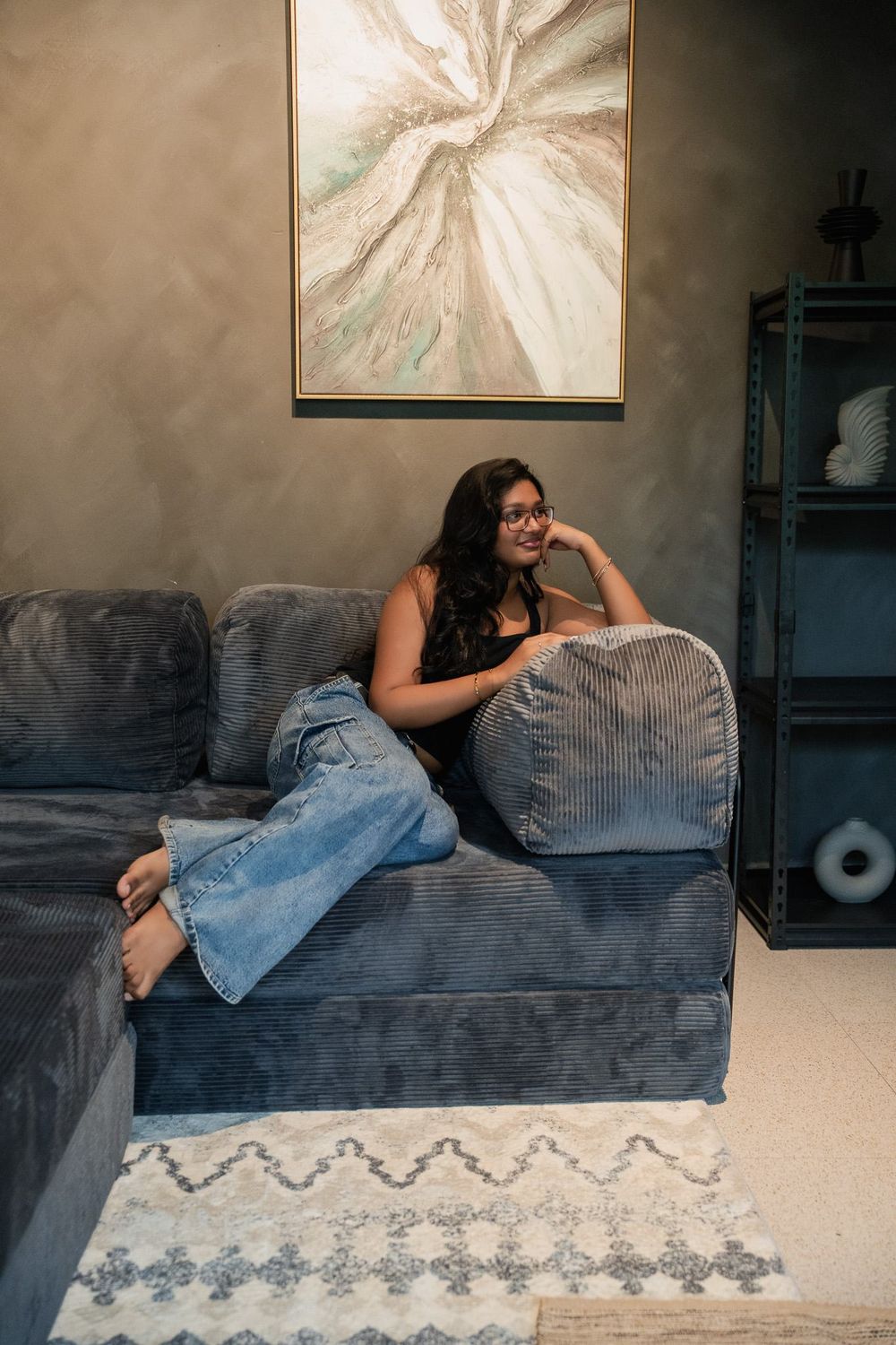 A woman reclining across the slate grey corduroy Cloud Corner, framed painting on the wall behind.